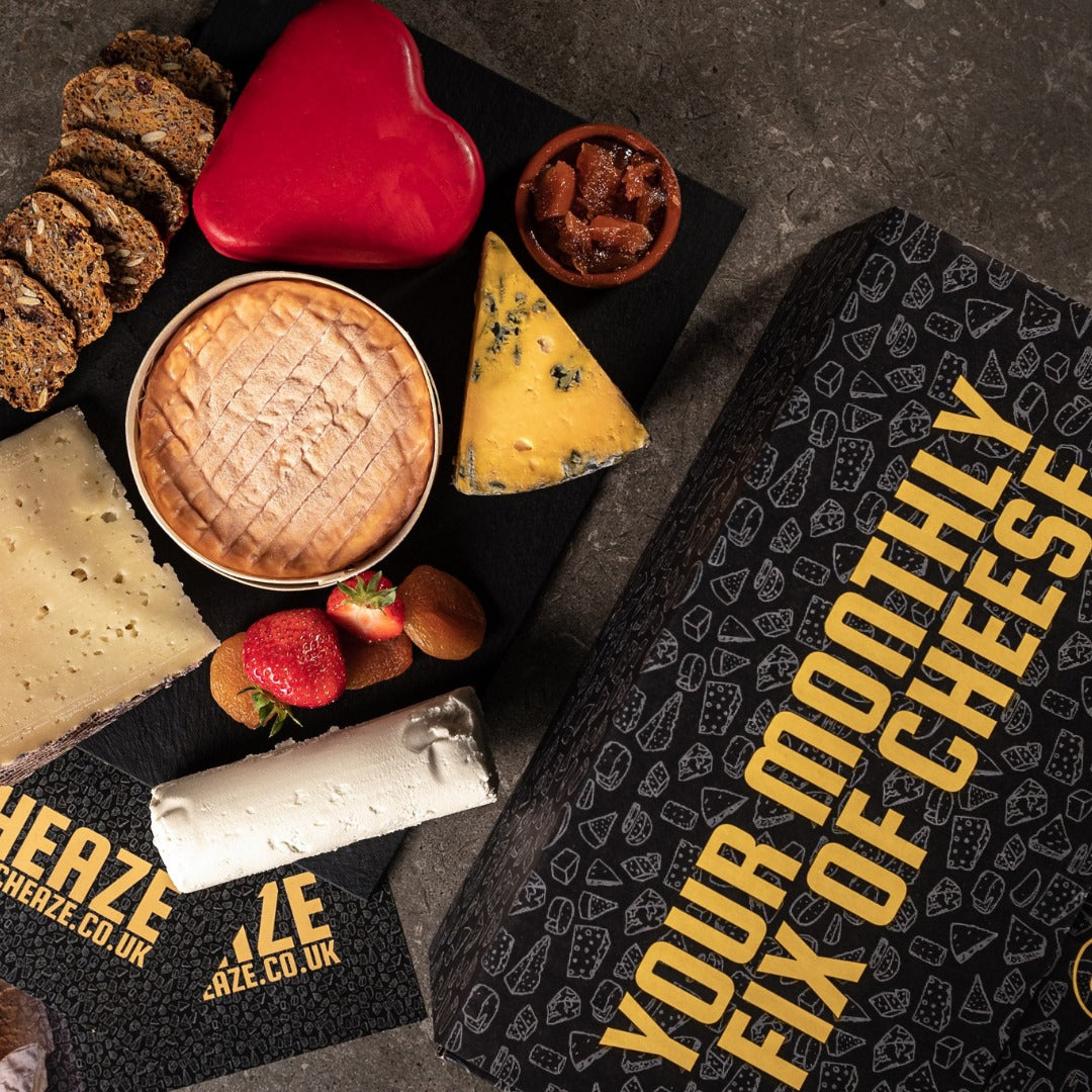 A red heart-shaped waxed cheese, a boxed soft cheese, blue and hard wedges, a white log and crackers beside a Cheaze cheese box