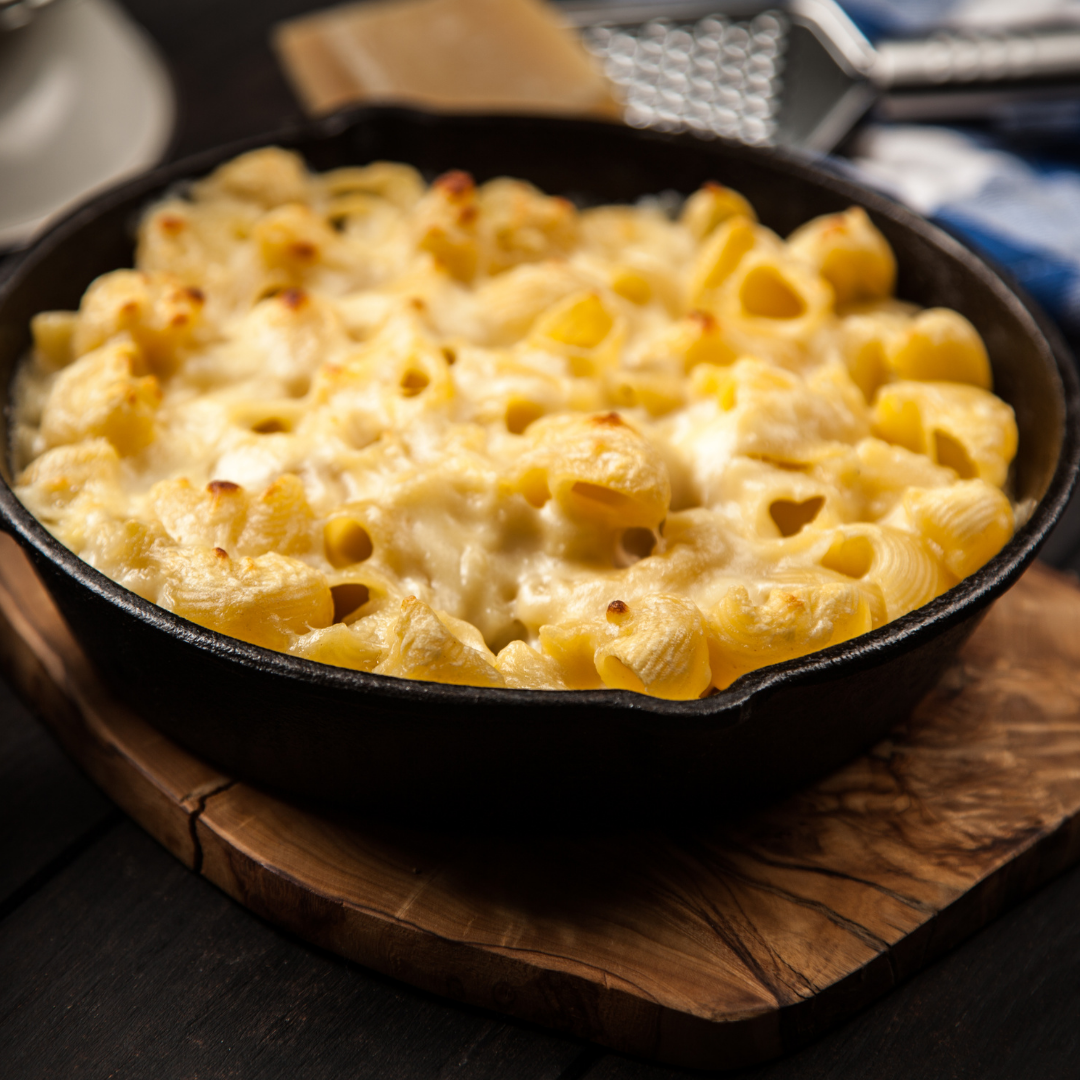 Baked macaroni cheese with a golden top in a cast iron skillet on a wooden board, a grater and cheese behind