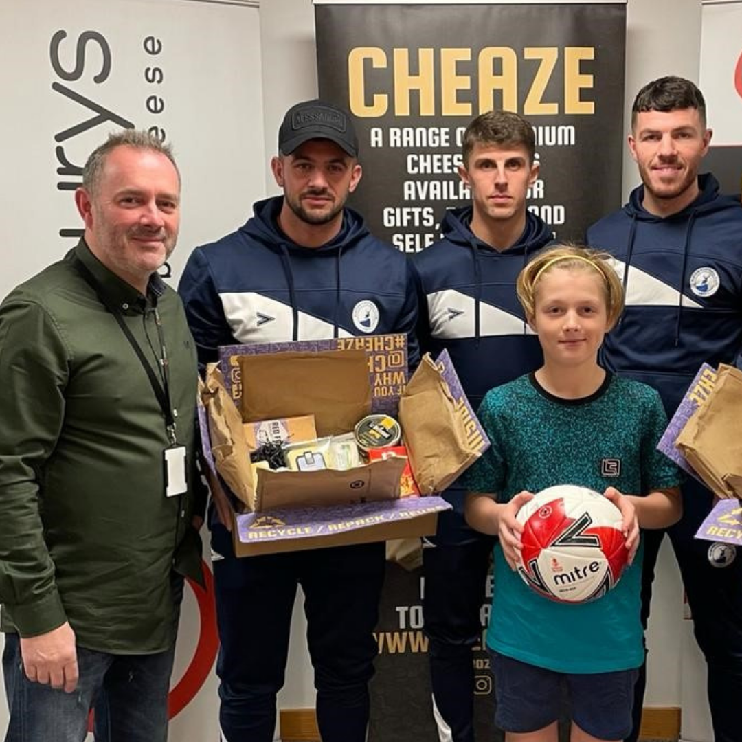 Buxton FC players in club tracksuits, one holding an open Cheaze cheese box, with a man and a boy holding a football