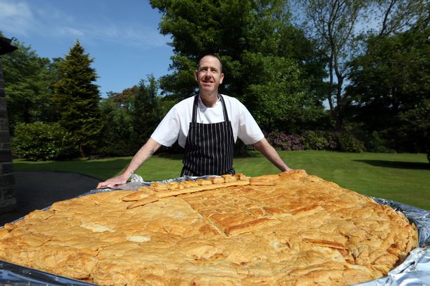 Chef in a striped apron behind the giant Noggie, a pasty made with Northumberland Oak Smoked cheese, on a lawn