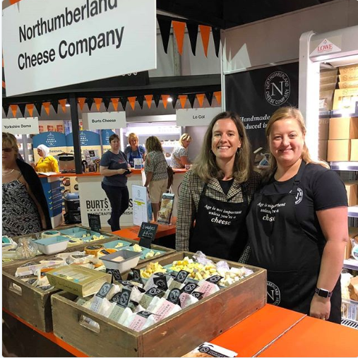 Two Northumberland Cheese Company team members in branded aprons at the company's show stand, behind crates of cheese