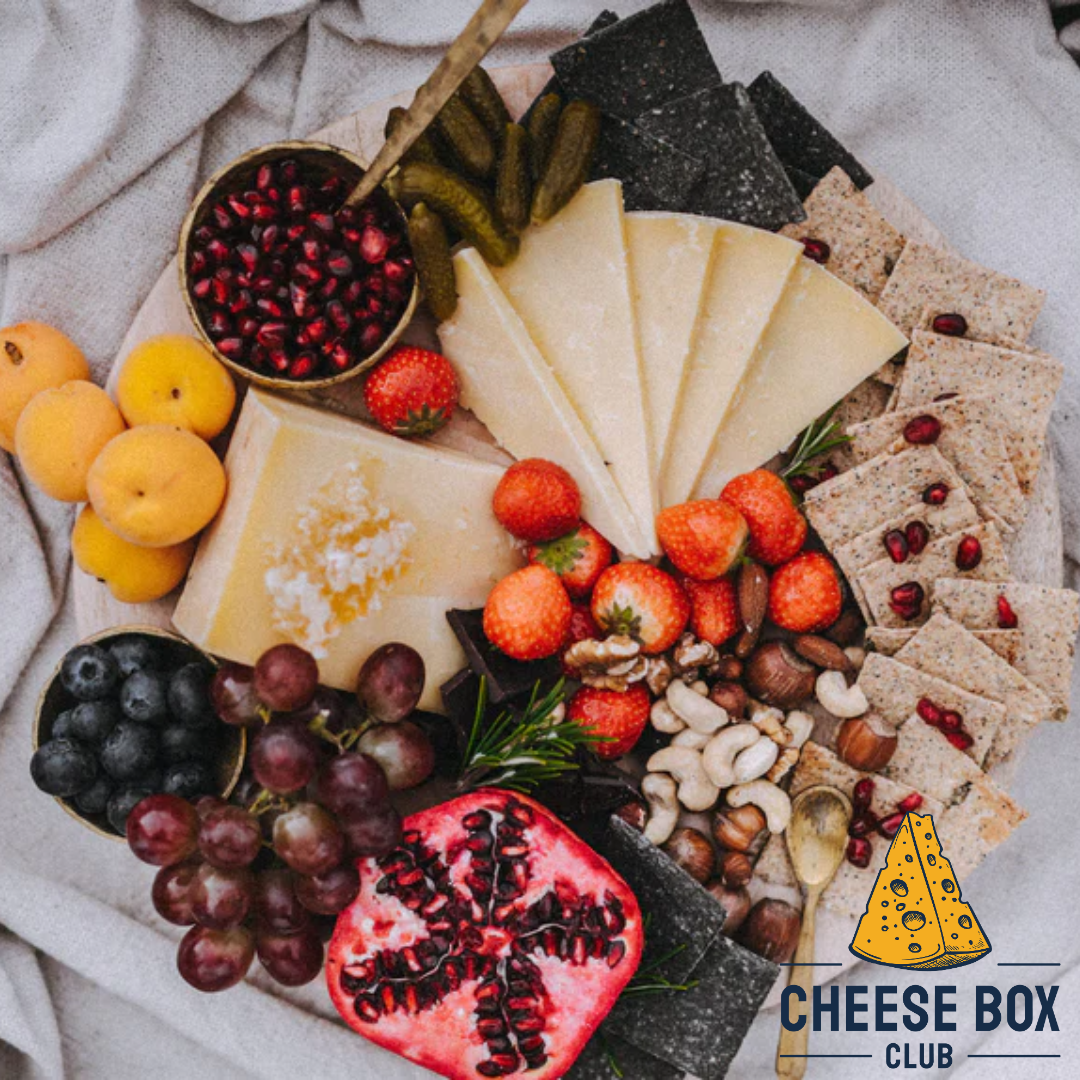 Cheeseboard of hard cheese wedges with strawberries, grapes, pomegranate, nuts and crackers, with the Cheese Box Club logo