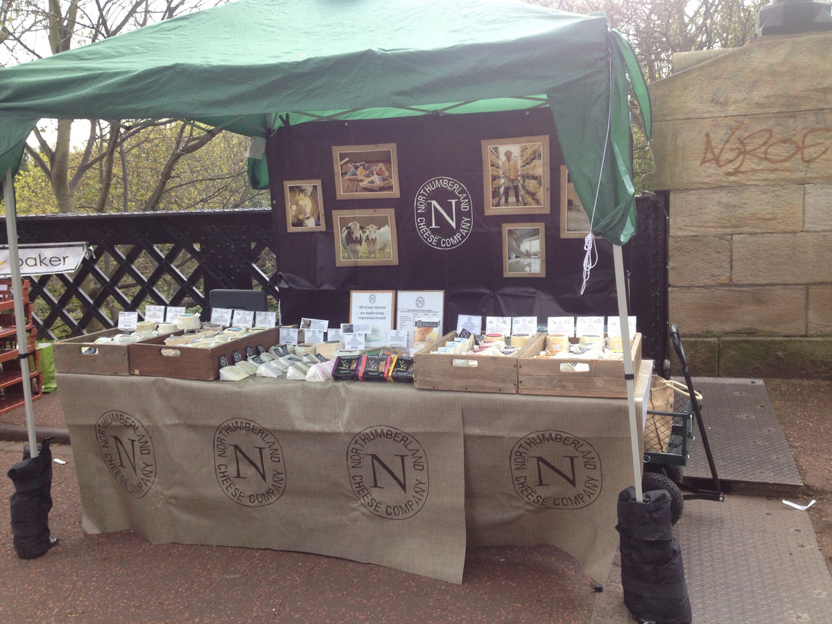 The Northumberland Cheese Company market stall under a green gazebo, cheeses in wooden crates on a branded hessian cloth