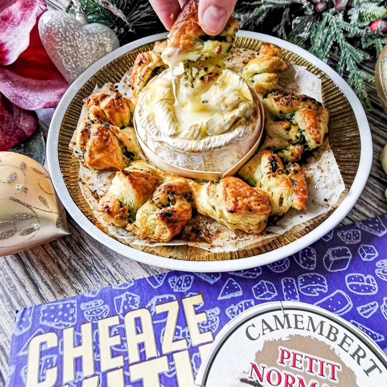 Hand dipping a pesto puff pastry twist into a baked Petit Normand Camembert ringed with twists on a gold platter