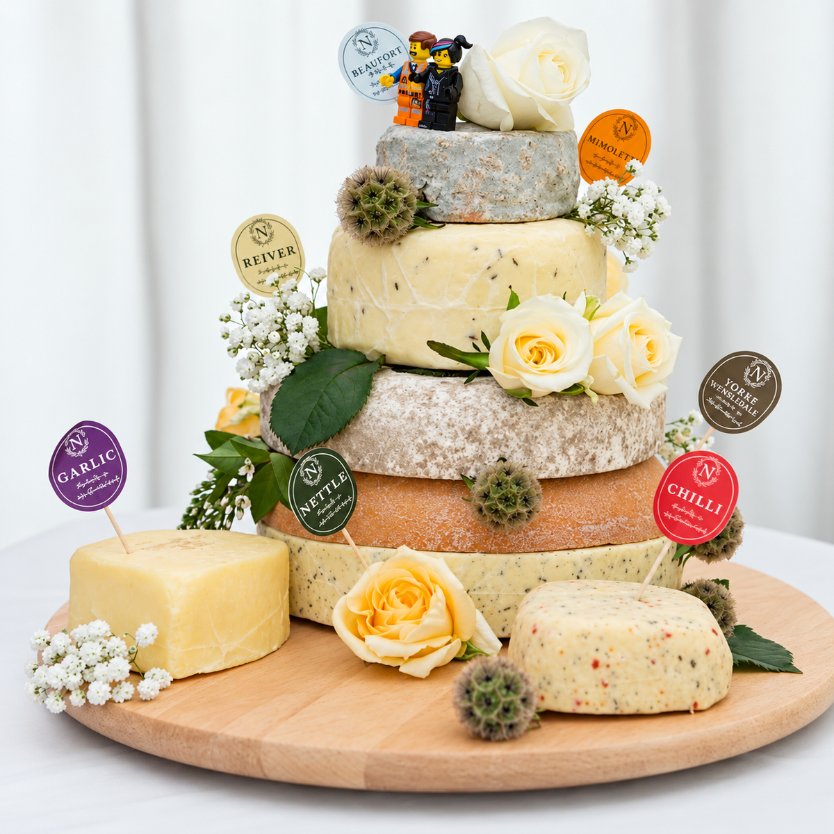Five-tier cheese wedding cake on a wooden board, decorated with white and yellow roses, gypsophila and two toy figures