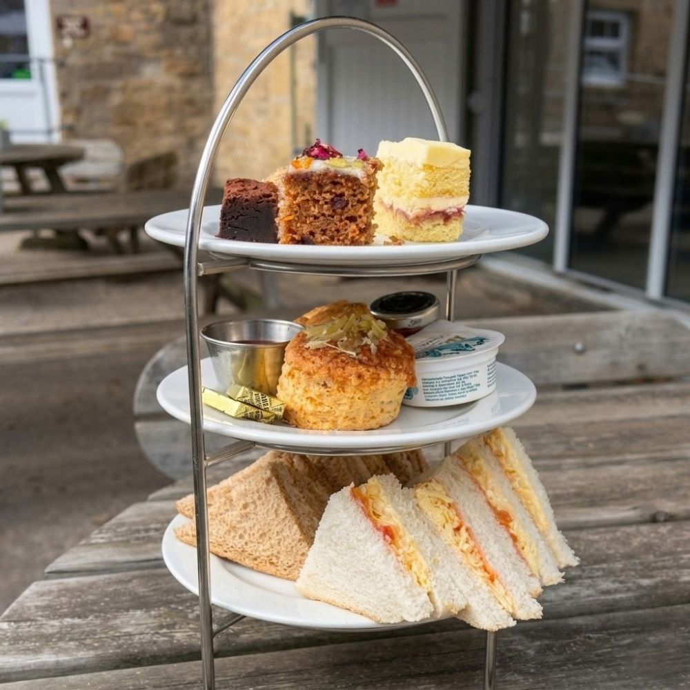 Afternoon tea on a three-tier stand: cheese and tomato sandwiches, a cheese scone with butter and jam, and slices of brownie, carrot cake and sponge on an outdoor table