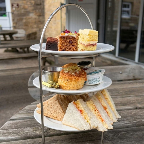 Afternoon tea on a three-tier stand: cheese and tomato sandwiches, a cheese scone with butter and jam, and slices of brownie, carrot cake and sponge on an outdoor table