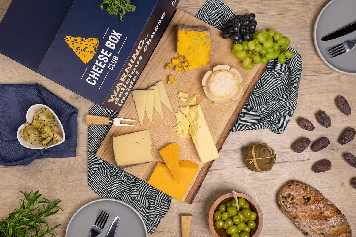 Navy Cheese Box Club box beside a board of artisan cheeses with grapes, olives, dates and a rustic loaf