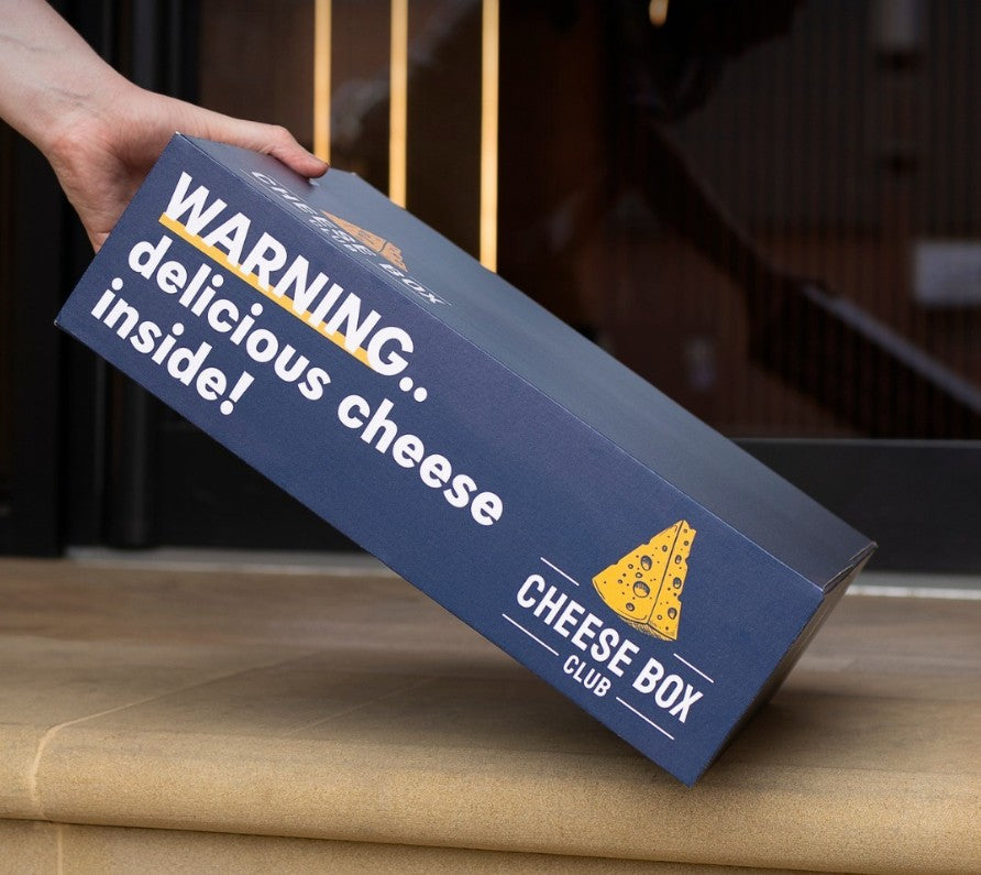 A navy Cheese Box Club box printed 'Warning.. delicious cheese inside!' being set down on a doorstep