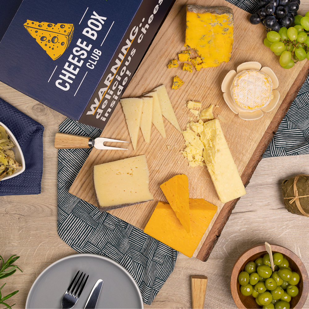 A navy Cheese Box Club box reading 'Warning: delicious cheese inside' beside a board of cheeses, grapes and olives