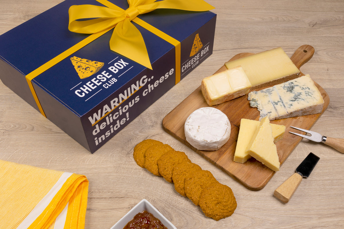Navy Cheese Box Club box with a yellow ribbon and the words delicious cheese inside, beside a board of cheeses and crackers