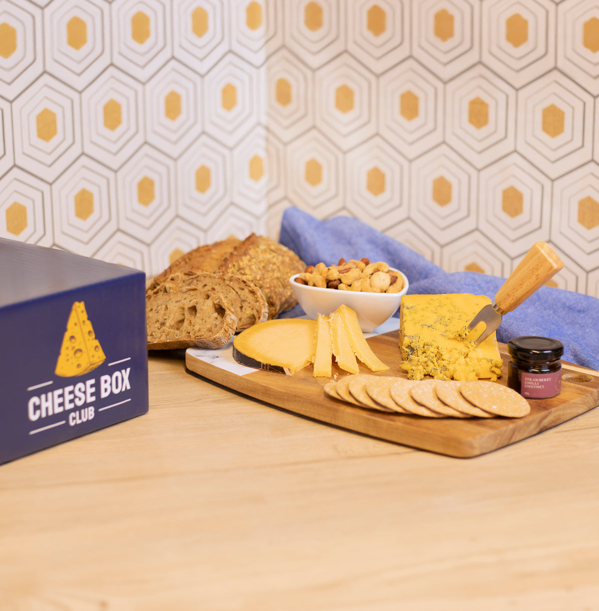 Navy Cheese Box Club box beside a board of cheeses, crackers and a small jar of chutney, with sliced bread and a bowl of nuts