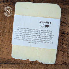 EweMoo Cheese block in its white paper label with sheep and cow symbols, laid on a dark rustic wooden table