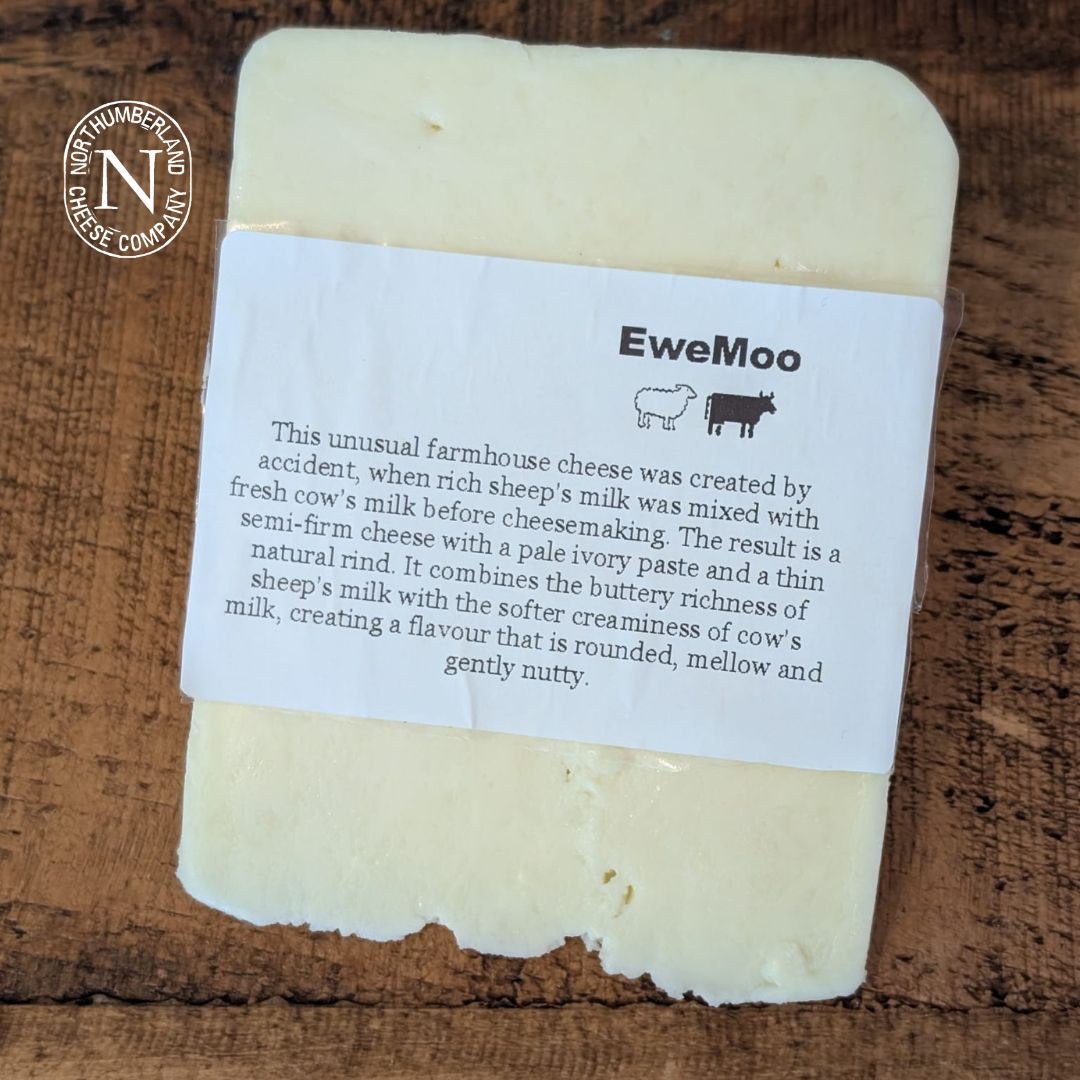EweMoo Cheese block in its white paper label with sheep and cow symbols, laid on a dark rustic wooden table