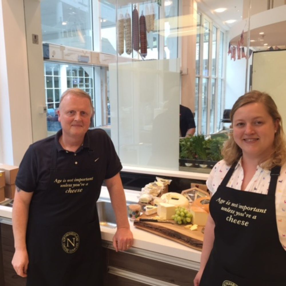 Two Northumberland Cheese Company team members in branded black aprons beside a board of cheeses at a deli counter