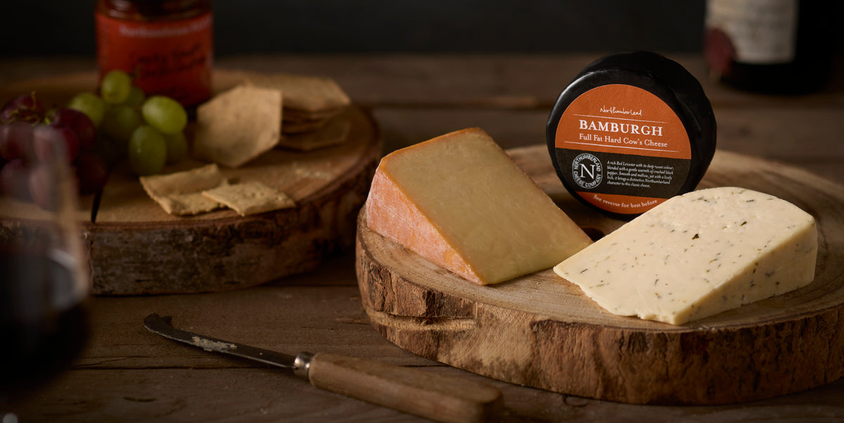 A Bamburgh truckle with wedges of Oak Smoked and Nettle cheese on a wooden board, with crackers, grapes and red wine
