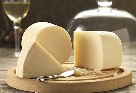 A whole round and cut wedges of pale sheep's milk cheese on a wooden board with a cheese knife and a glass of white wine