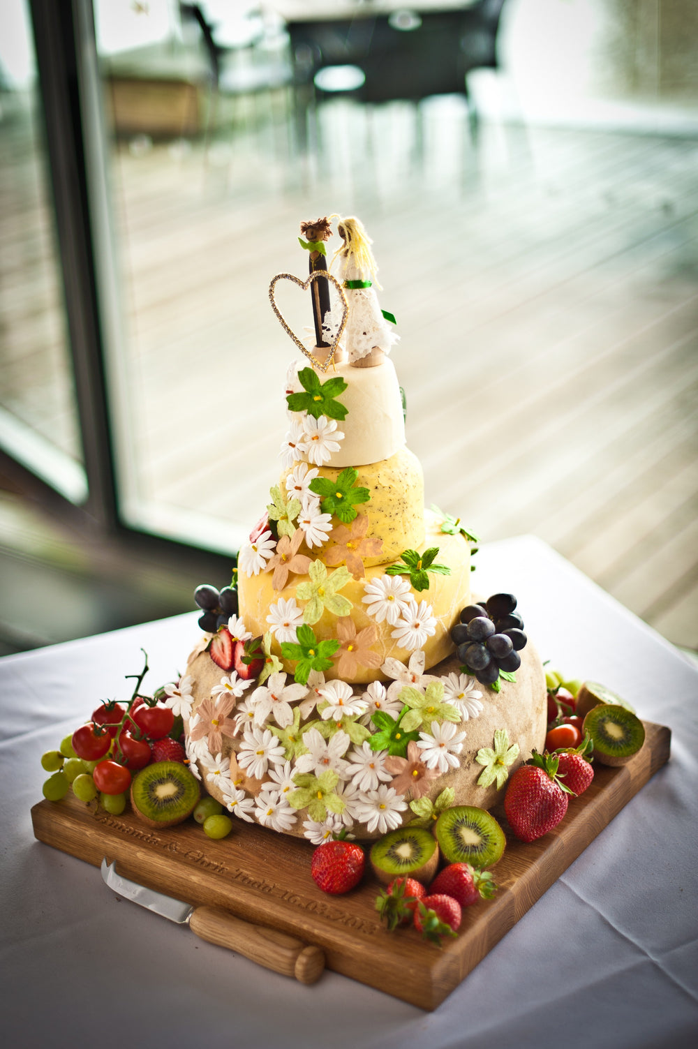 Four-tier cheese wedding cake dressed with daisies and a bride and groom topper, surrounded by grapes, strawberries and kiwi