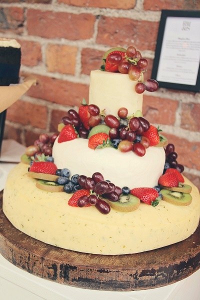 Three-tier cheese wedding cake topped with strawberries, grapes, blueberries and kiwi slices on a wooden board