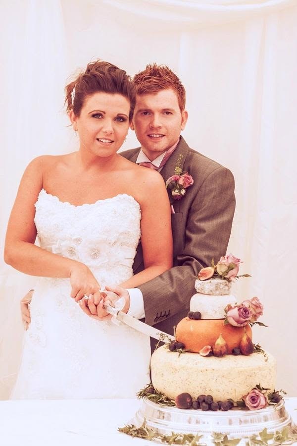 Bride and groom cutting their tiered cheese wedding cake, decorated with pink roses and figs