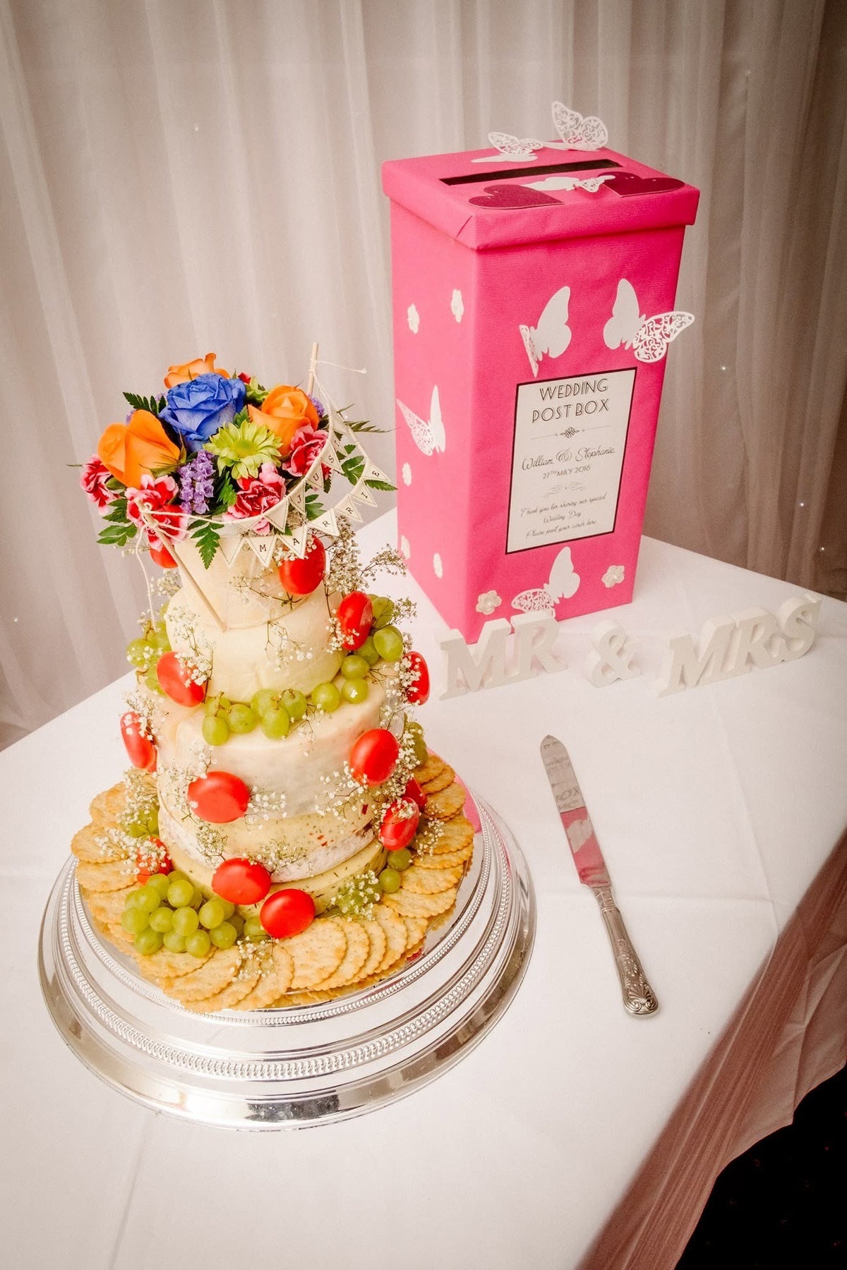 Cheese wedding cake topped with bright flowers and bunting, with grapes and crackers, on a silver stand by a wedding post box