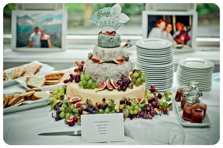 Tiered cheese wedding cake with figs and grapes under a Say Cheese topper, on a buffet table with crackers and chutneys