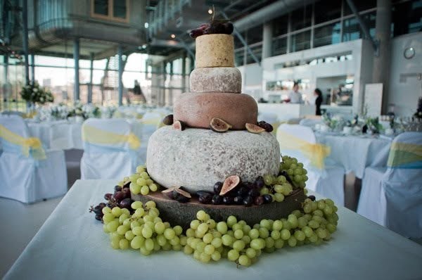 Tiered cheese wedding cake with figs, ringed with green and red grapes, in a glass-walled room set for a wedding reception