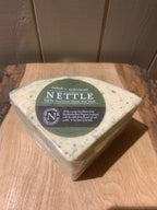 Triangular wedge of Nettle Cheese flecked with nettles, wrapped in clear film with its green label, on a rustic wooden board