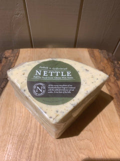 Triangular wedge of Nettle Cheese flecked with nettles, wrapped in clear film with its green label, on a rustic wooden board