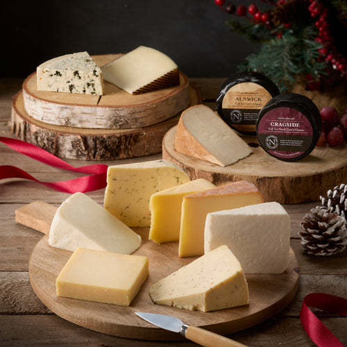 Twelve Cheeses of Christmas Box: seven cheese wedges on a board; on log slices a russet-rinded wedge, the Alnwick and Cragside truckles, a blue cheese and a Manchego