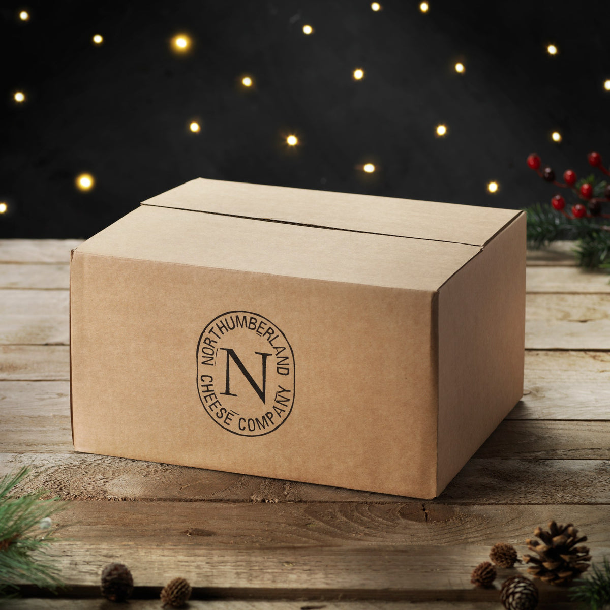 Brown box for the Twelve Cheeses of Christmas Box, stamped with the Northumberland Cheese Company logo, among fairy lights and pine cones